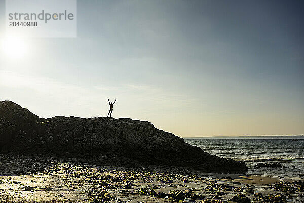 Man with arms raised standing on rock near sea at sunrise