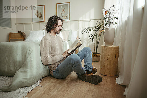 Handsome man reading book sitting by bed at home