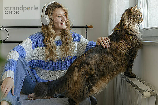 Happy woman wearing wireless headphones and sitting near cat looking through window at home