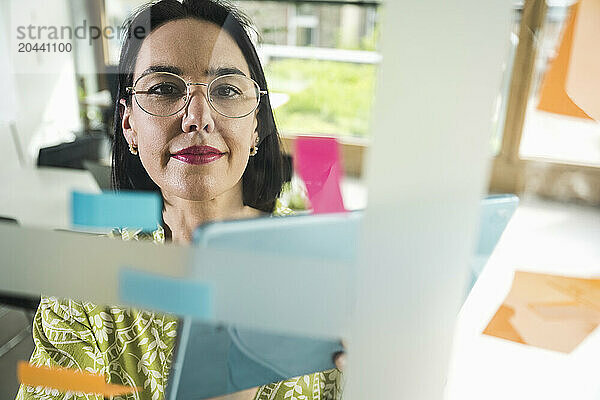 Confident mature businesswoman sticking adhesive notes on glass in office