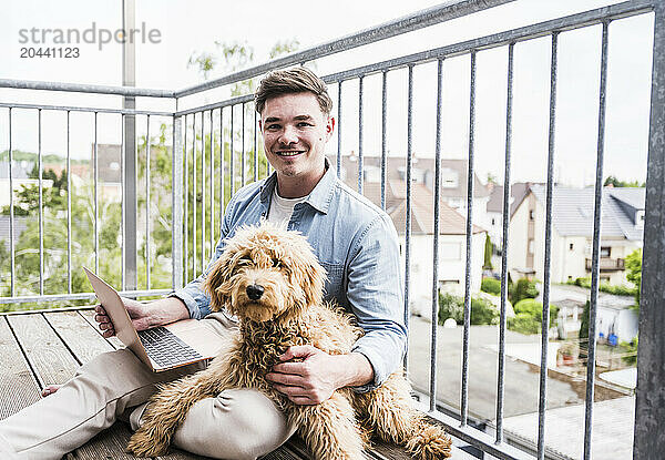 Happy man sitting with dog and laptop on balcony