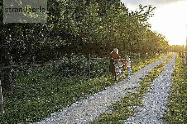 Mother and daughter walking with bicycle on footpath at sunset