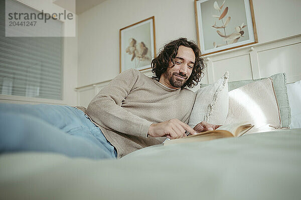 Smiling man reading book reclining on bed at home