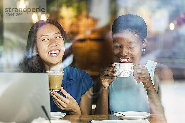 Happy multiracial friends drinking coffee sitting with laptop at table in cafe