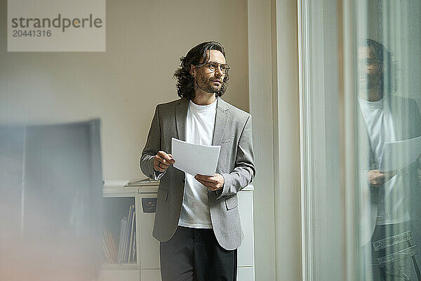 Mature businessman standing with document looking out through window at office