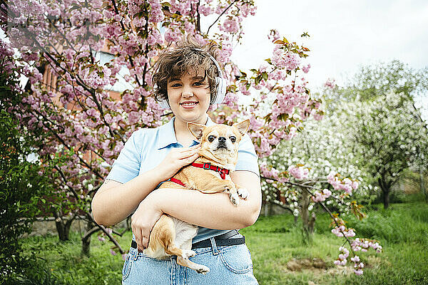 Smiling woman listening to music through wireless headphones carrying dog in garden