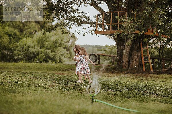 Siblings running near garden hose splashing water on grass