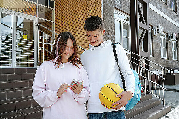 Teenage girl sharing smart phone with boy standing at schoolyard