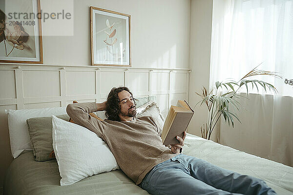 Handsome man reading book lying on bed at home