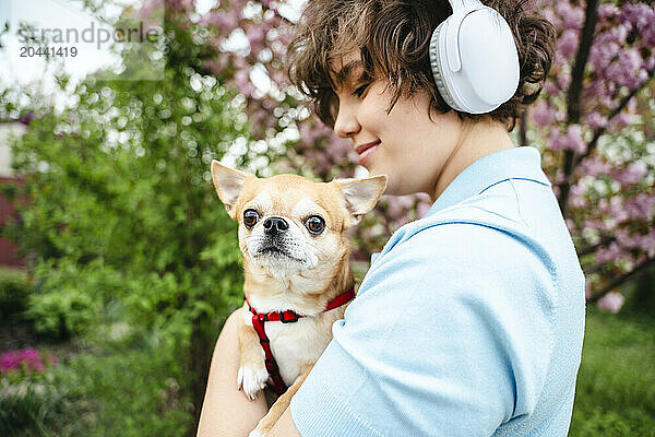 Young woman listening to music through wireless headphones carrying dog in garden