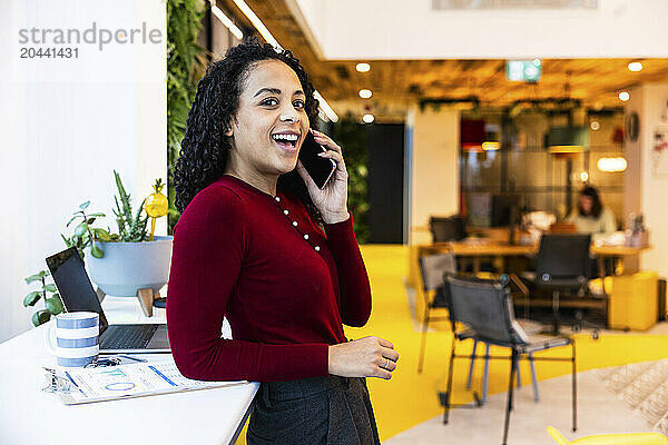 Happy businesswoman talking on smart phone leaning at desk in office