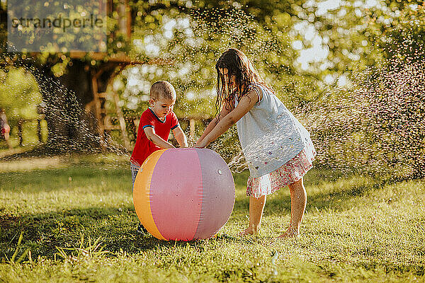 Brother and sister dancing by colorful ball splashing water at garden