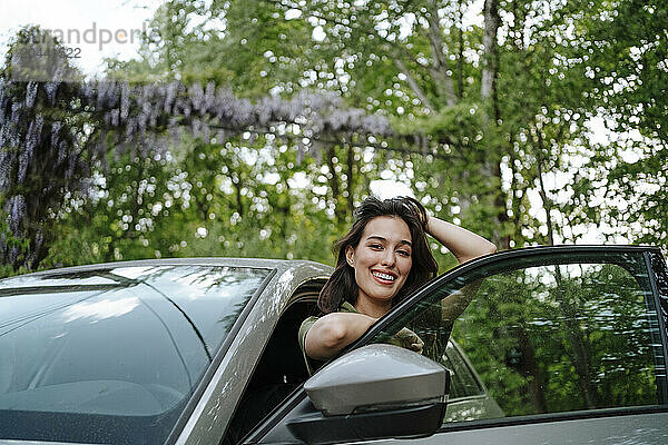 Smiling young woman standing with car in front of tree