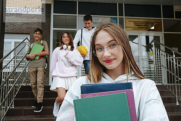 Smiling teenage girl wearing eyeglasses with books standing in front of friends at schoolyard