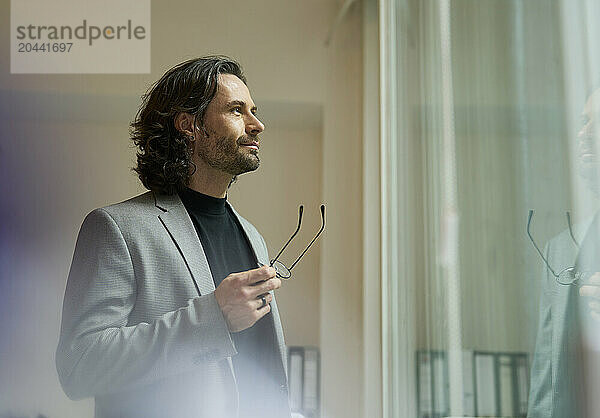 Mature businessman standing with eyeglasses looking out through window at office