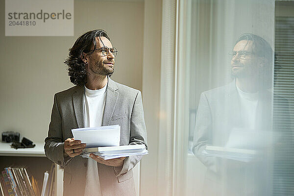 Handsome businessman standing with document looking out through window at office