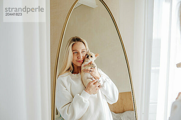 Smiling woman holding chihuahua dog and looking in mirror at home