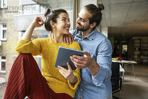 Smiling young couple with tablet PC looking at each other near window