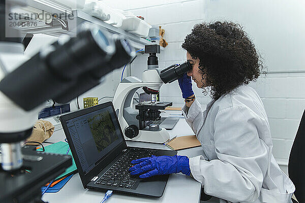 Young scientist using microscope sitting with laptop at laboratory