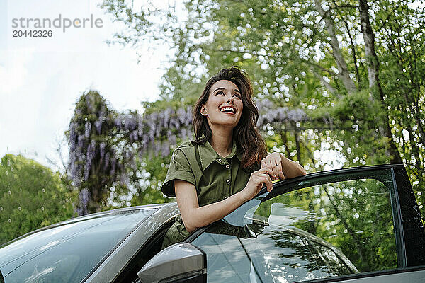 Happy woman leaning on car door in front of tree