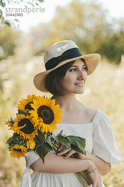 Thoughtful woman wearing hat and holding sunflowers at field