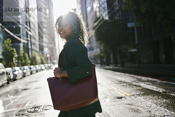 Back lit smiling young businesswoman with purse crossing street in city