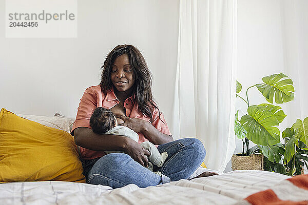 Mother breastfeeding daughter and sitting cross-legged on bed at home