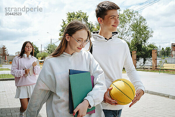 Smiling teenage girl and boy walking together with friend in background at schoolyard