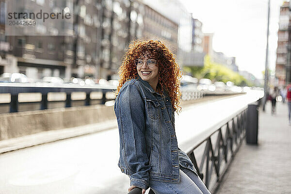 Smiling redhead woman with curly hair sitting on railing in city