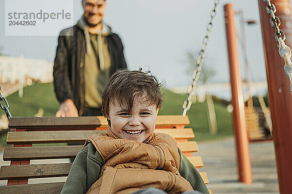 Smiling son sitting on swing with father at public park