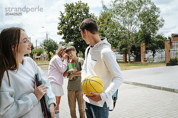 Teenage girl whispering to boy standing near friends at schoolyard on sunny day