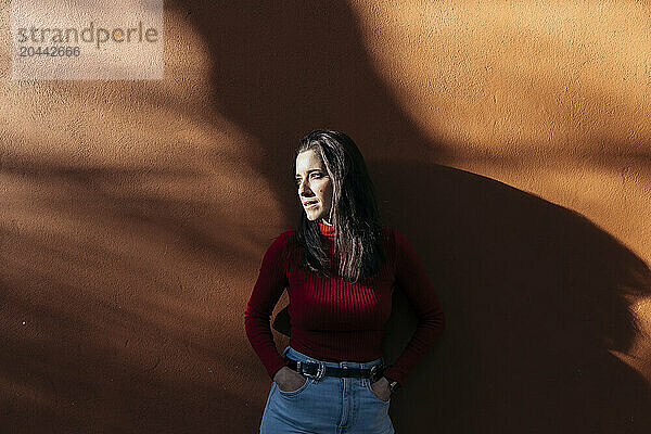 Thoughtful woman with hands in pockets standing near wall