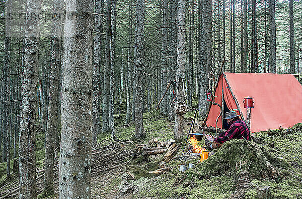 Senior man enjoying bonfire and cooking food near tent in forest