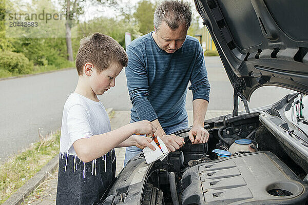 Boy using dipstick by father leaning on car