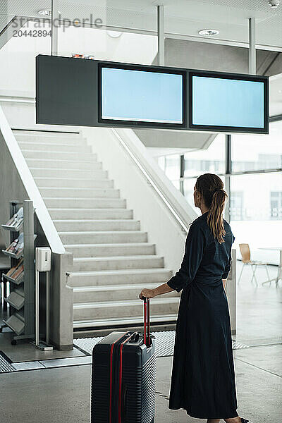 Businesswoman holding suitcase and looking at arrival departure board