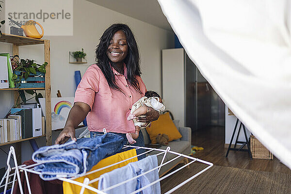 Happy woman keeping clothes on drying rack and holding daughter at home