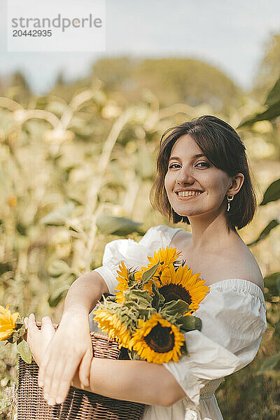 Smiling woman holding sunflowers basket at field