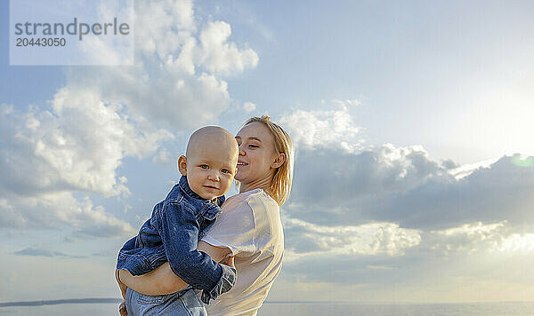 Smiling mother holding baby in arms under cloudy sky