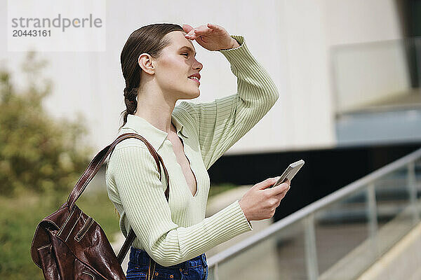 Woman holding smartphone and shielding eyes near railing