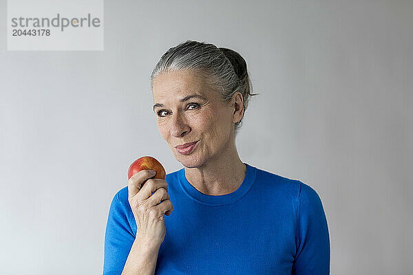 Smiling senior woman holding apple in front of wall