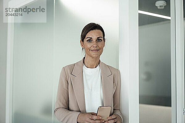 Smiling business professional holding smart phone and standing near wall at office