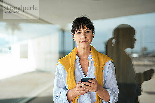 Confident businesswoman holding mobile phone in front of glass wall