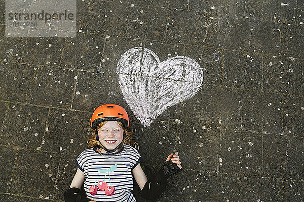 Smiling girl wearing helmet lying down near heart shape drawing on ground