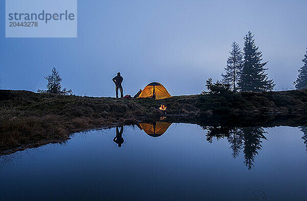 Senior man standing near tent and camping at lake shore