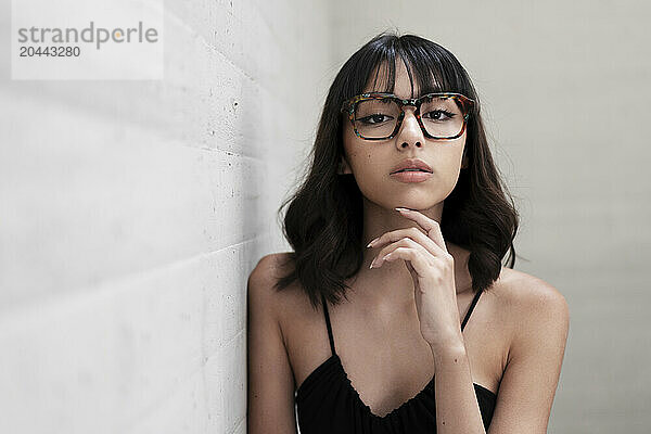 Confident young woman wearing eyeglasses leaning on gray wall