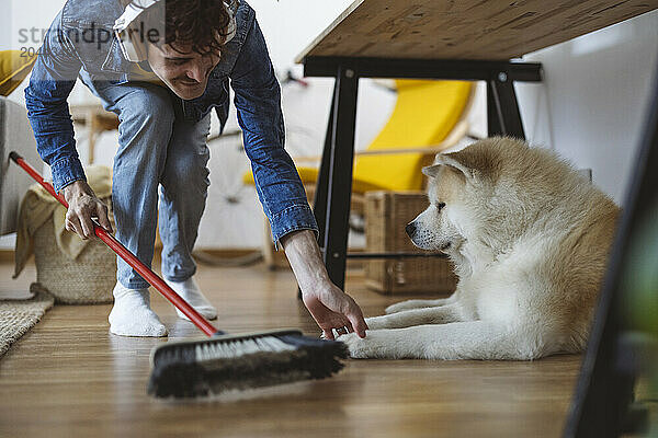 Smiling man wearing wireless headphones and sweeping floor near dog at home