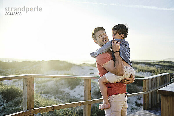 Father carrying son by boardwalk railing at beach