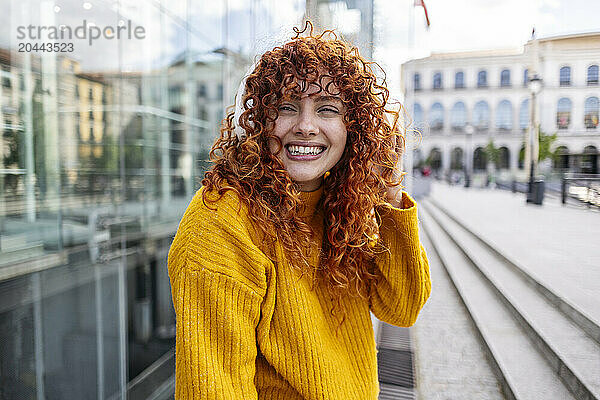 Happy redhead woman listening to music through wireless headphones in city