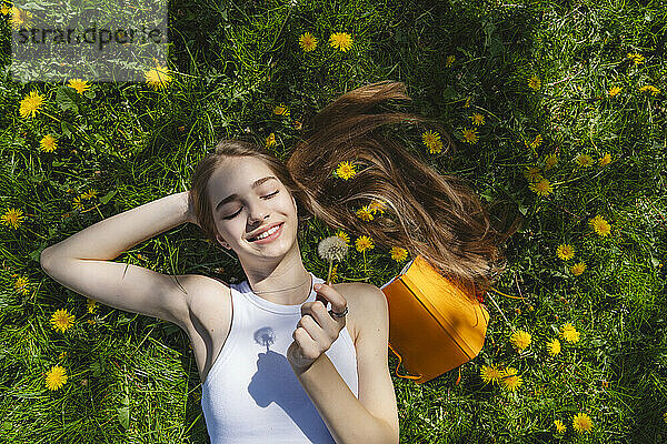 Smiling teenage girl lying with yellow book over dandelions in park