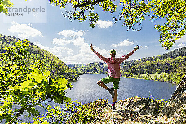 Woman with arms raised standing on rock in front of Obersee lake at north Rhine Westphalia  Eifel national park  Germany